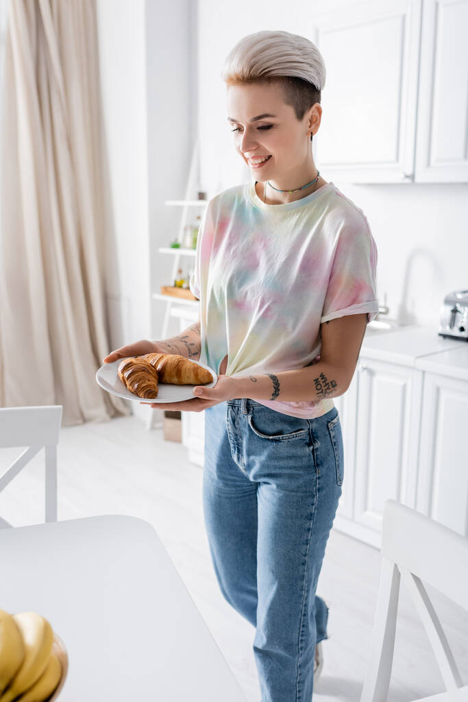 smiling tattooed woman in jeans holding plate with delicious croissants in kitchen - Fotó, kép