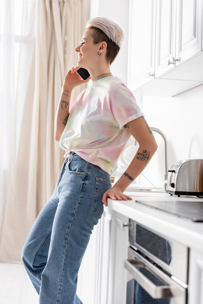 вид сбоку счастливой женщины в джинсах, стоящей на кухне и разговаривающей по мобильному телефону - Фото, изображение