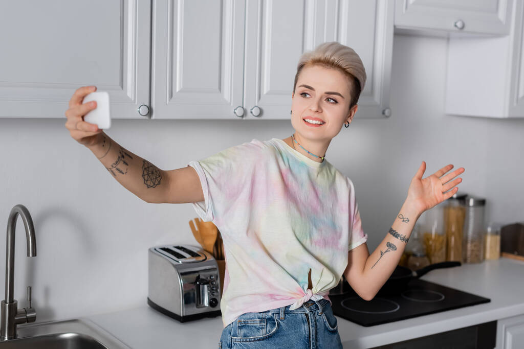 smiling tattooed woman taking selfie on cellphone in kitchen - Photo, image