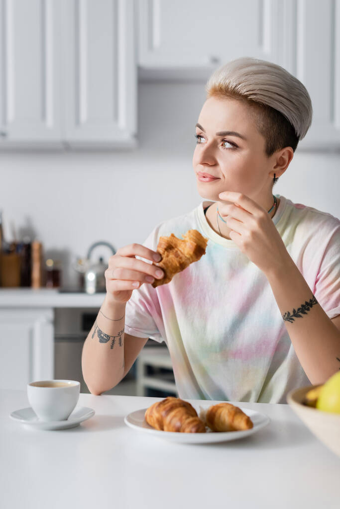 positive woman looking away while sitting with croissant near coffee cup - Фото, изображение