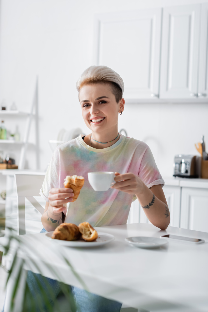 vidám nő kávéscsészével és finom croissant nézi kamera a konyhában - Fotó, kép