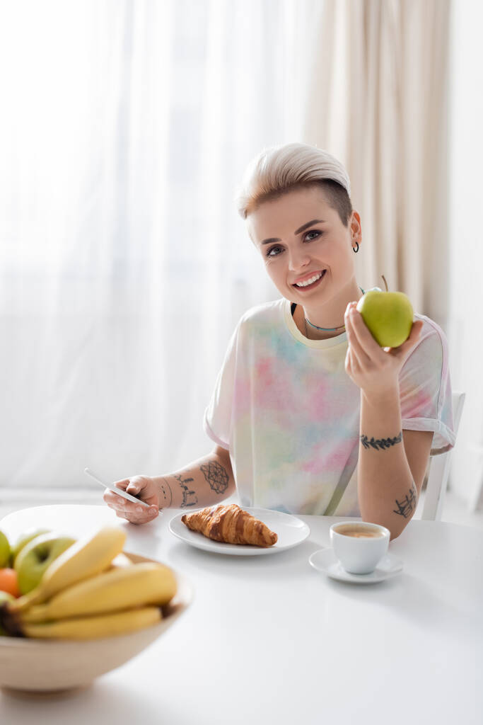 happy and stylish woman holding apple and mobile phone near plate with croissant - Фото, изображение