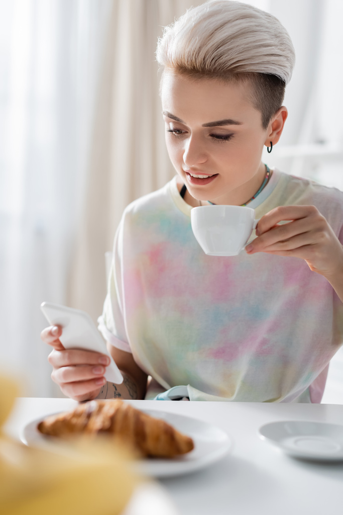 uśmiechnięta kobieta z filiżanką kawy przy użyciu smartfona w pobliżu rogalika na niewyraźnym pierwszym planie - Zdjęcie, obraz