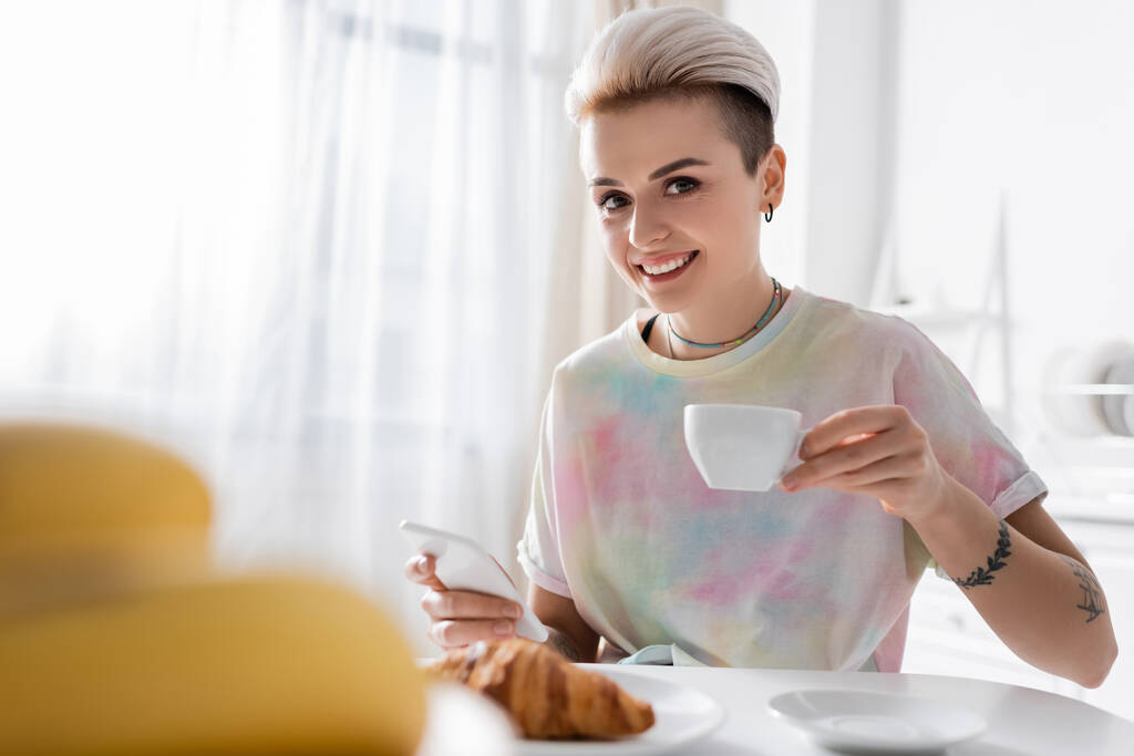 stílusos nő okostelefonnal és kávéscsészével mosolygós kamera homályos előtérben - Fotó, kép