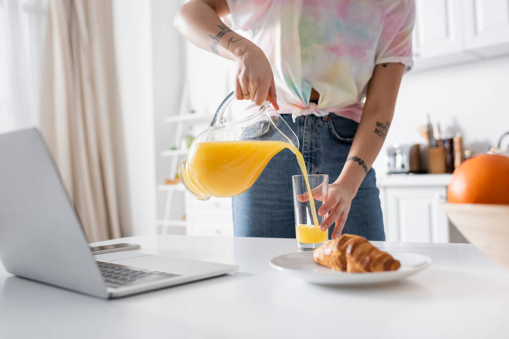 cropped view of tattooed woman pouring orange juice near croissant and blurred laptop - 写真・画像