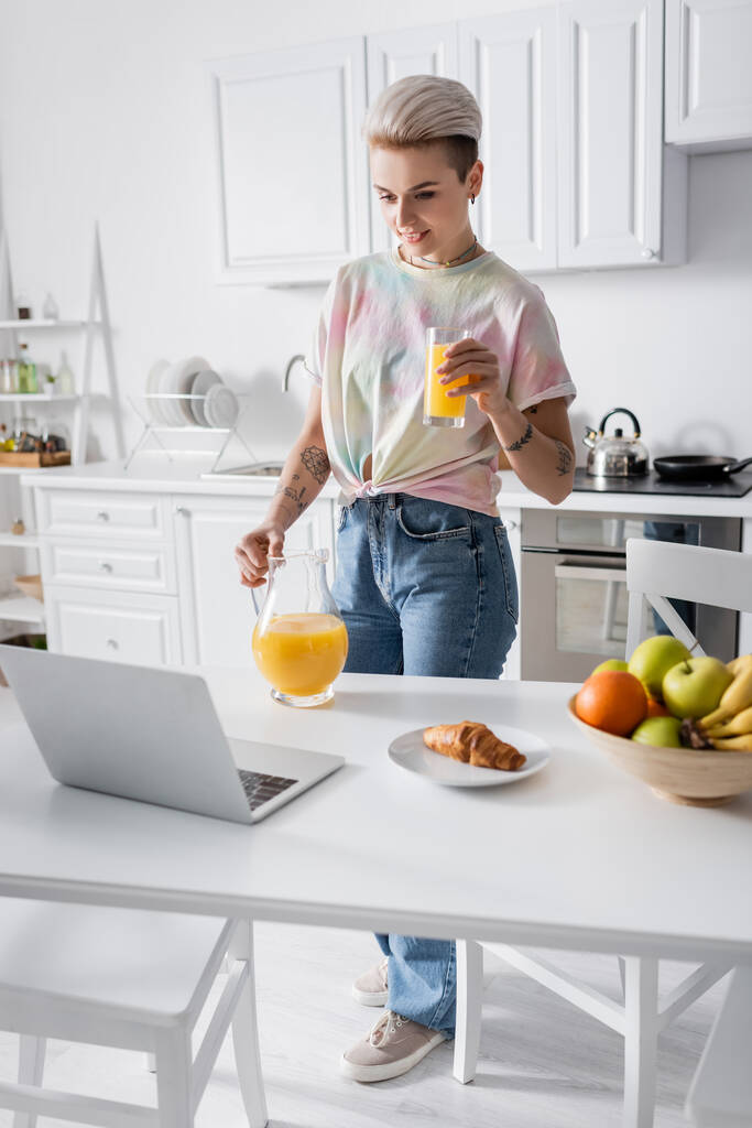 молода жінка з апельсиновим соком біля ноутбука, круасану та свіжих фруктів на кухонному столі
 - Фото, зображення