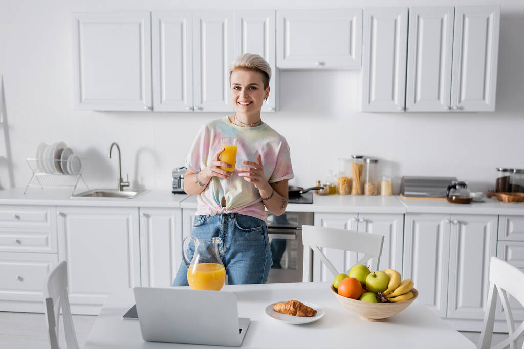 trendy woman with orange juice smiling at camera near kitchen table with laptop and breakfast - Foto, Imagem