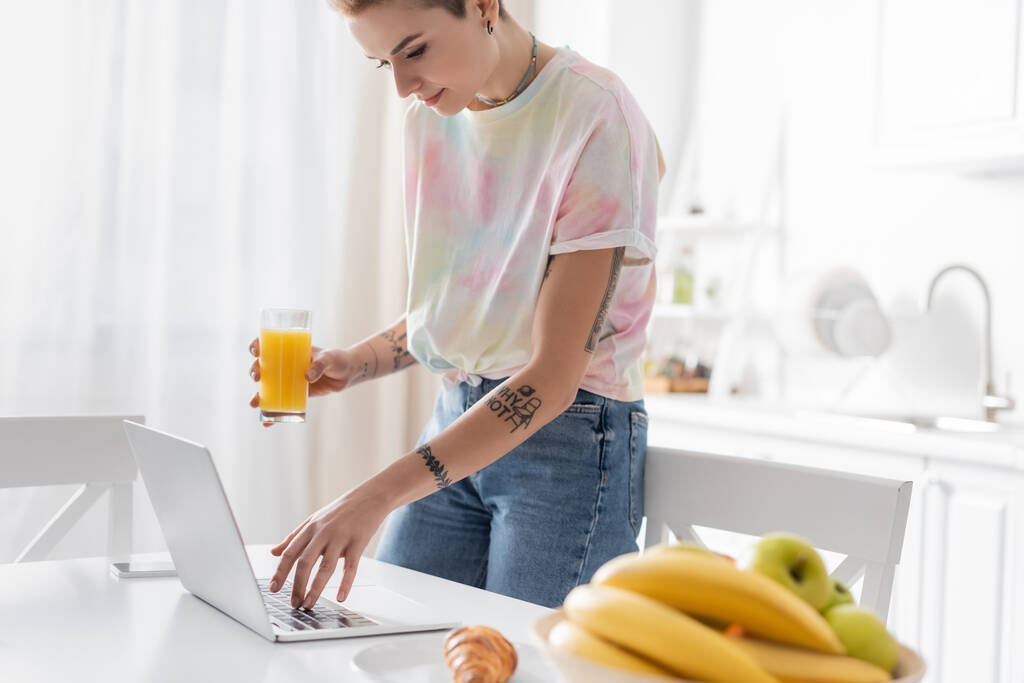 jeune femme avec du jus d'orange en utilisant un ordinateur portable près croissant et des fruits flous dans la cuisine - Photo, image