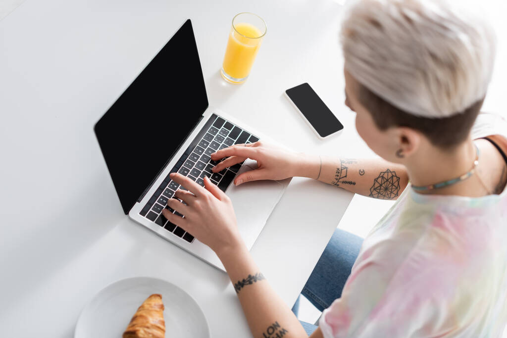 overhead view of blurred woman typing on laptop near smartphone, orange juice and croissant - Fotó, kép