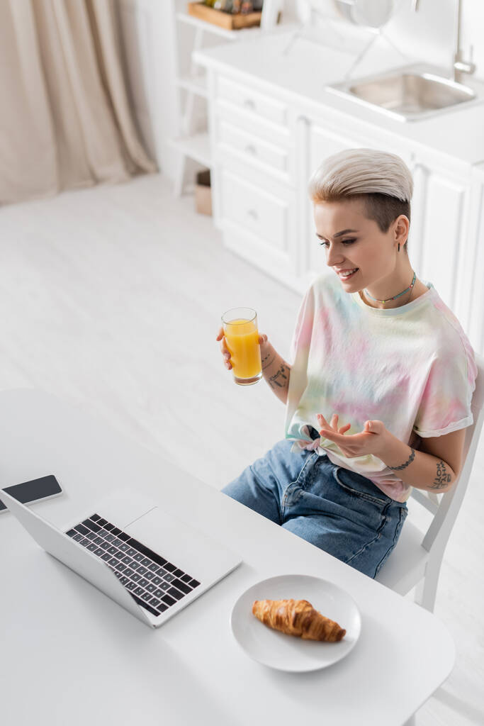 smiling woman with orange juice pointing at laptop near croissant and smartphone with blank screen - Photo, image