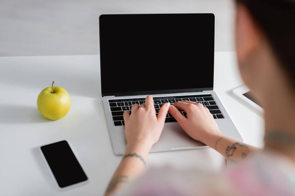 vista recortada de la mujer borrosa escribiendo en el ordenador portátil cerca de teléfono inteligente con pantalla en blanco y manzana madura - Foto, imagen