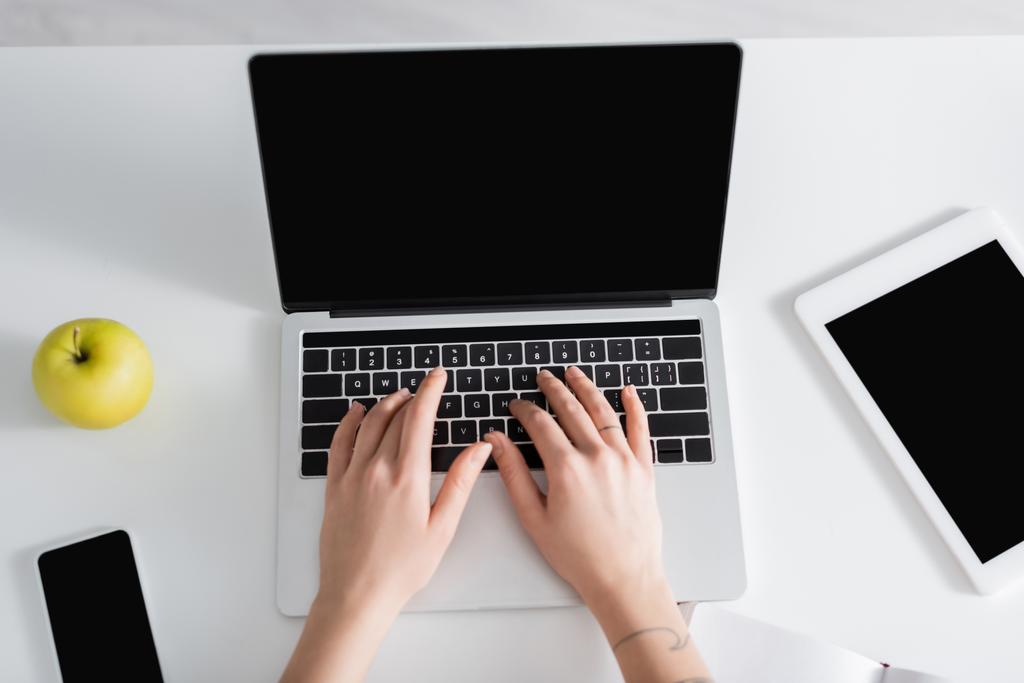 вид сверху женщины, печатающей на ноутбуке рядом со смартфоном, цифровым планшетом и свежим яблоком - Фото, изображение