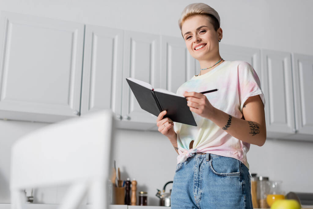 nízký úhel pohledu šťastné ženy s notebookem a perem při pohledu do kamery v kuchyni - Fotografie, Obrázek