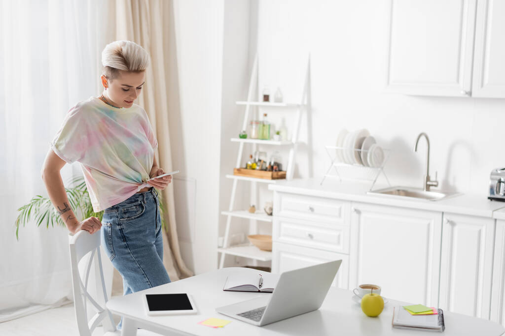 stylish woman standing with mobile phone near laptop, digital tablet and notebooks on kitchen table - Foto, Imagem