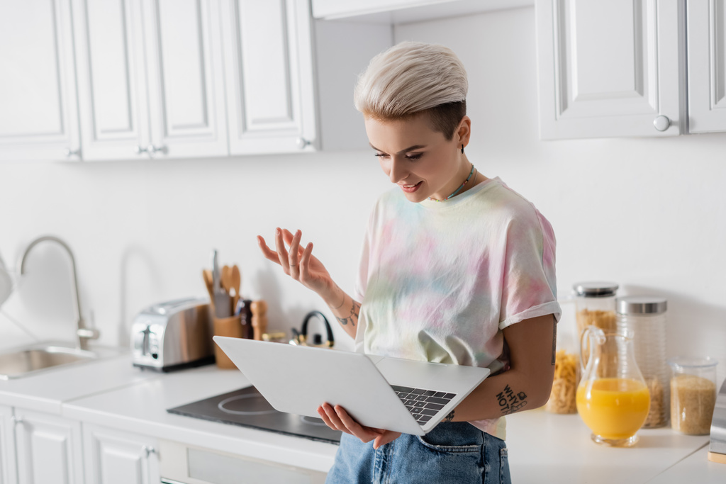 trendy getatoeëerde vrouw op zoek naar laptop en gebaren in de keuken - Foto, afbeelding