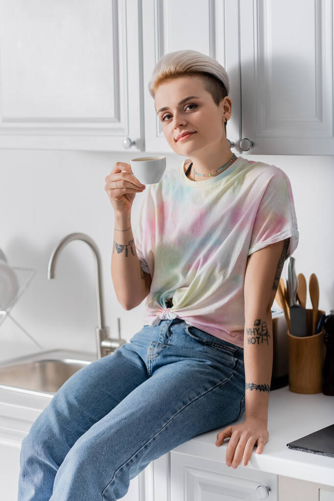 smiling woman sitting on kitchen worktop with coffee cup and looking at camera - Foto, Imagem
