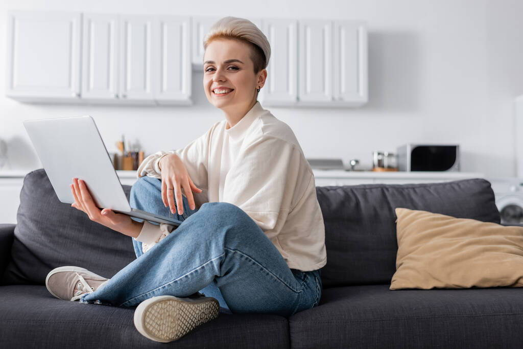 woman with laptop sitting on couch with crossed legs and smiling at camera - Photo, Image