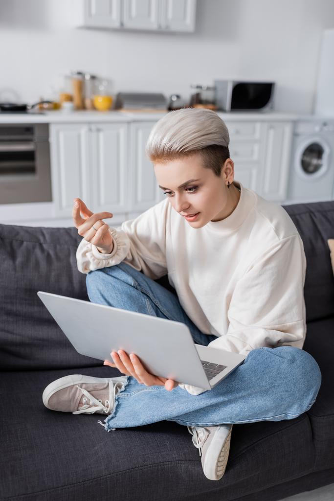trendy woman sitting on couch with crossed legs during video call on laptop - Zdjęcie, obraz
