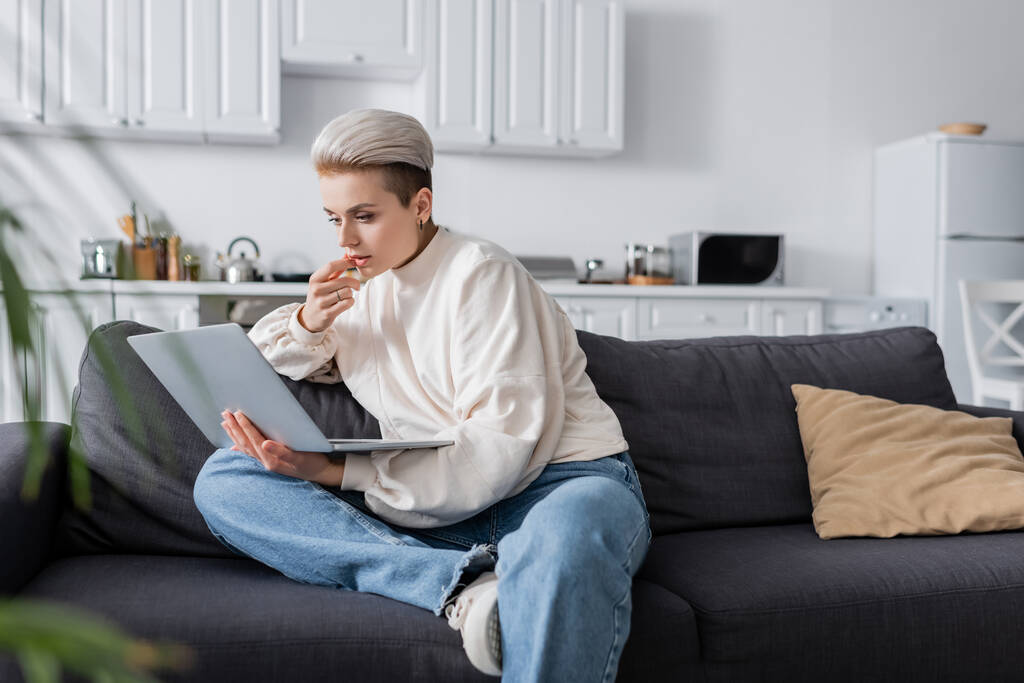 Beyaz kazaklı düşünceli bir kadın kanepeye oturmuş dizüstü bilgisayarına bakıyor. - Fotoğraf, Görsel