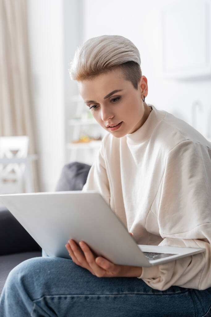 νέα και κομψή γυναίκα κοιτάζοντας φορητό υπολογιστή, ενώ κάθεται στο σπίτι - Φωτογραφία, εικόνα