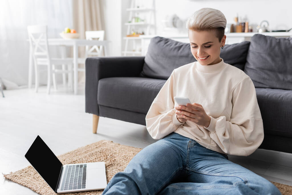 happy woman sitting on floor with mobile phone near laptop with blank screen - Foto, imagen