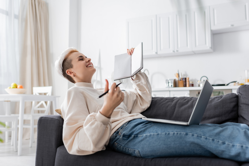 excited woman with blank notebook laughing near laptop on couch - Foto, Imagem