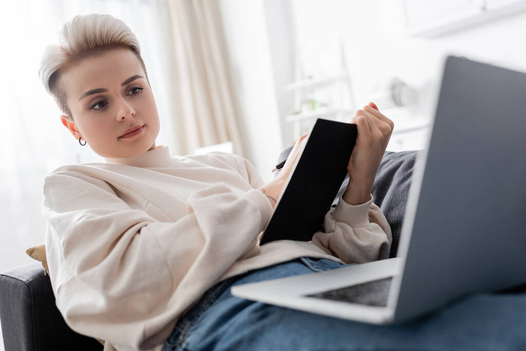 kobieta z modnej fryzury pisania w notatniku na kanapie w pobliżu zamazanego laptopa - Zdjęcie, obraz