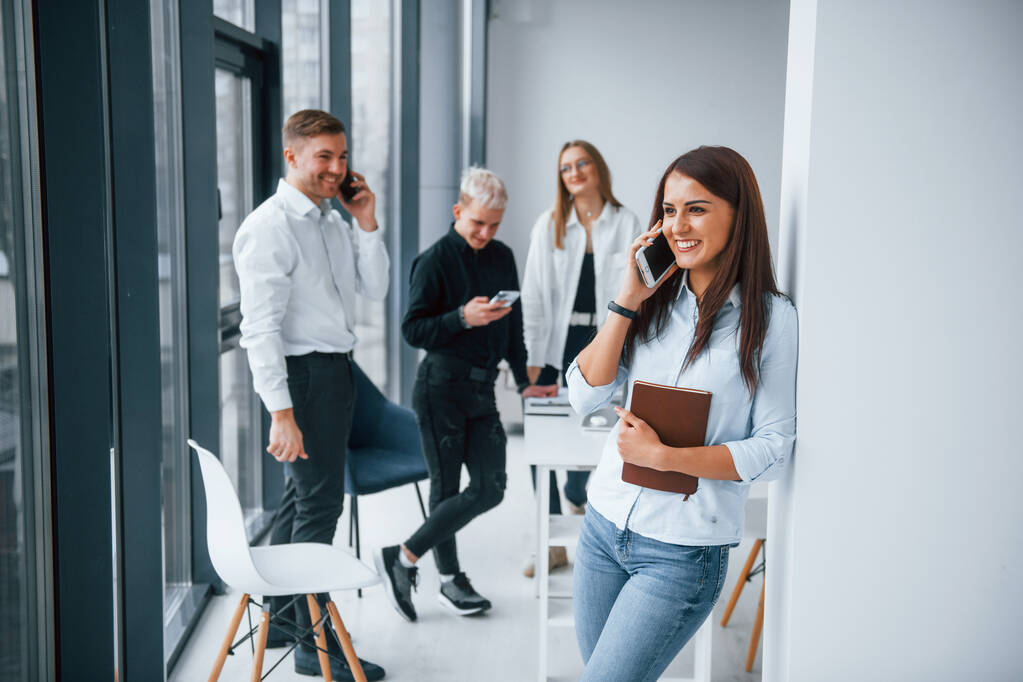 Egy vidám nő portréja egy csésze itallal és telefonnal, ami fiatal sikeres csapat előtt áll, akik együtt dolgoznak és kommunikálnak az irodában.. - Fotó, kép