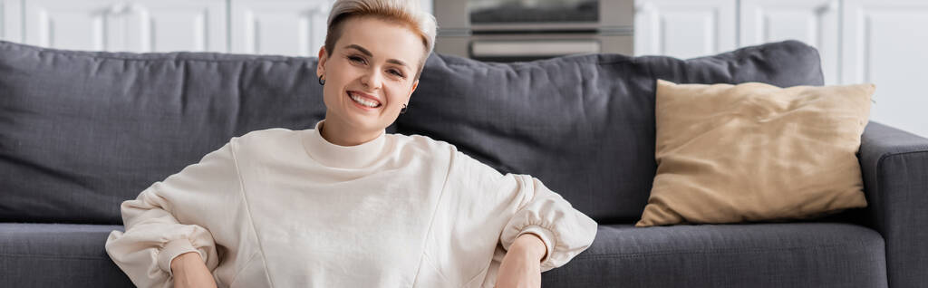 happy woman in white pullover smiling at camera near couch and pillow, banner - Photo, Image