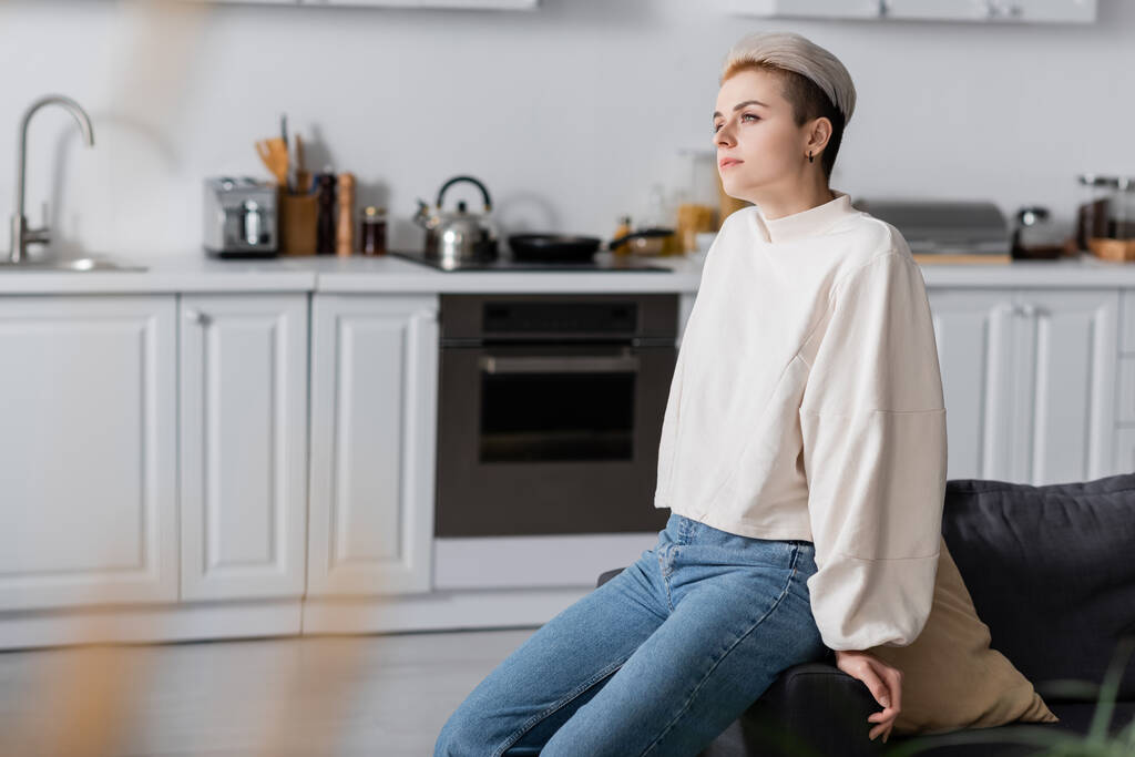 dreamy woman in white pullover sitting in kitchen and looking away - Foto, Bild