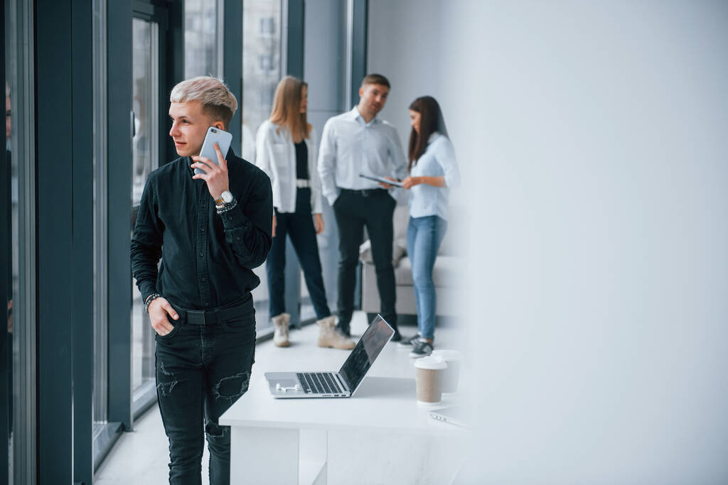 Νέοι hipster με τηλέφωνο στέκεται μπροστά από την ομάδα των νέων επιτυχημένη ομάδα που εργάζονται και επικοινωνούν μαζί σε εσωτερικούς χώρους στο γραφείο. - Φωτογραφία, εικόνα