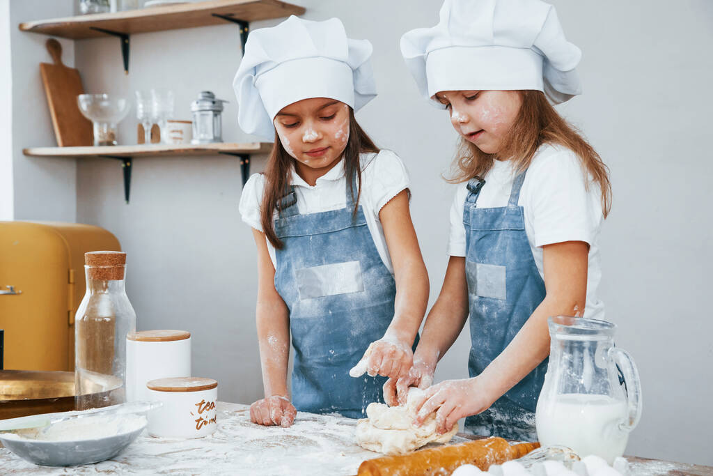 Дві маленькі дівчата в синьому шеф-кухарі рівномірно замішують тісто на кухні
. - Фото, зображення