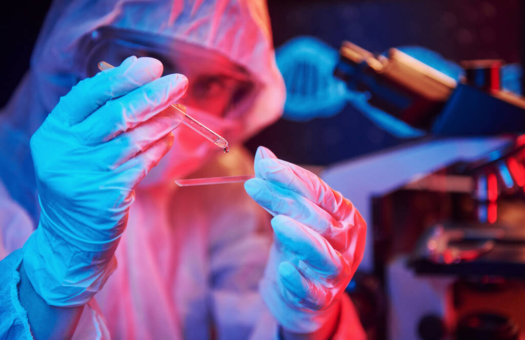 Infirmière masquée et uniforme blanc, tenant un tube avec liquide et assise dans un laboratoire éclairé au néon avec ordinateur et équipement médical à la recherche du vaccin contre le coronavirus. - Photo, image