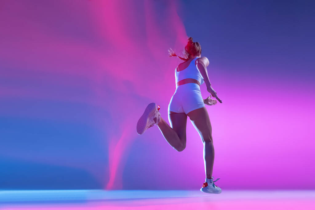 Zadní pohled. Profesionální atletka utíká izolované na modrém pozadí studia ve smíšeném růžovém neonovém světle. Zdravý životní styl, sport, pohyb a akční koncepce. - Fotografie, Obrázek