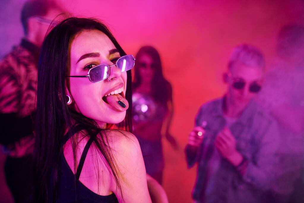 Jong meisje met drugs pillen op togue in het bijzijn van jongeren die plezier hebben in de nachtclub met kleurrijke laserlichten. - Foto, afbeelding