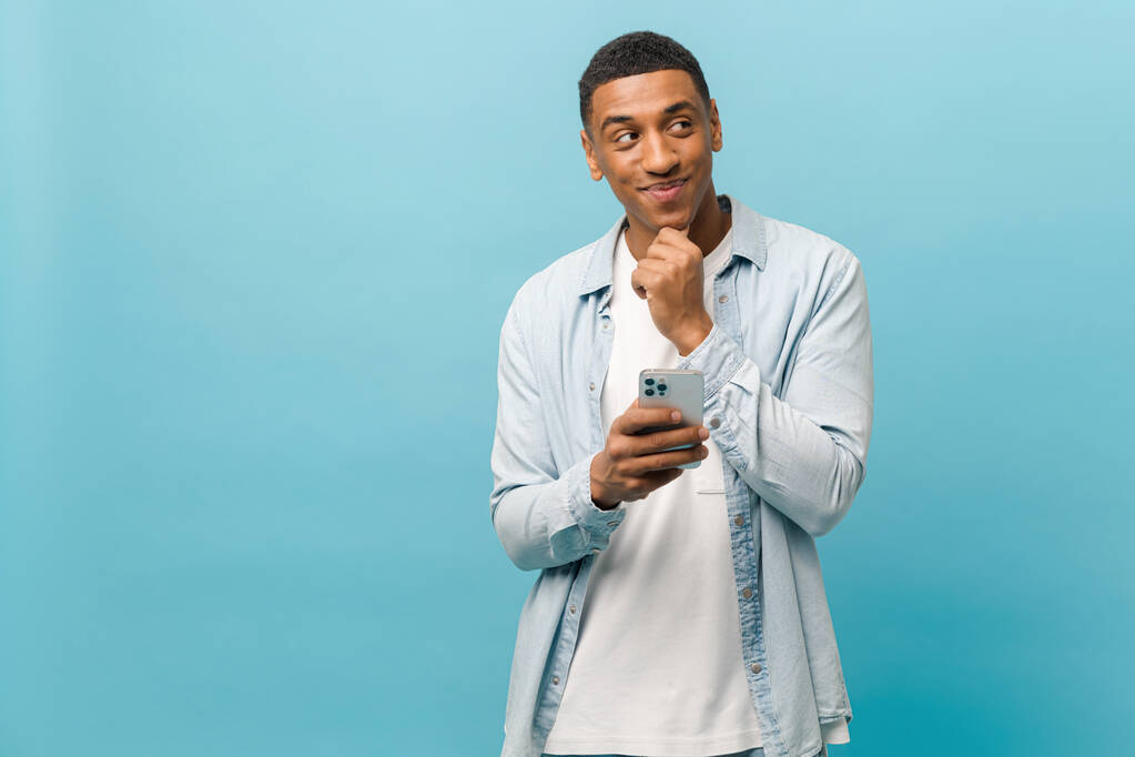 Задумчивый молодой многорасовый короткометражный мужчина в повседневной рубашке стоит со смартфоном и держит подбородок - Фото, изображение