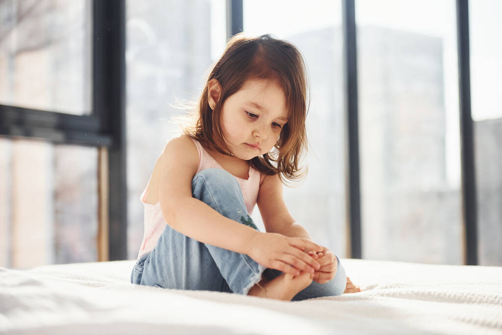 Cute little girl in casual clothes sits on bed alone at daytime. - Foto, immagini
