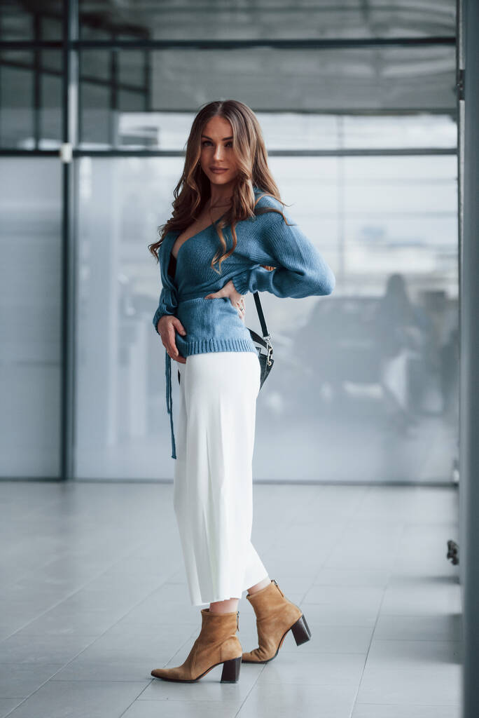 Jolie femme en chemise bleue et pantalon blanc à l'intérieur posant pour un appareil photo. À l'aéroport ou en magasin de voitures. - Photo, image