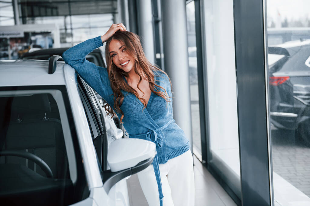 Positive woman in blue shirt leaning on brand new car. In auto salon. - Valokuva, kuva