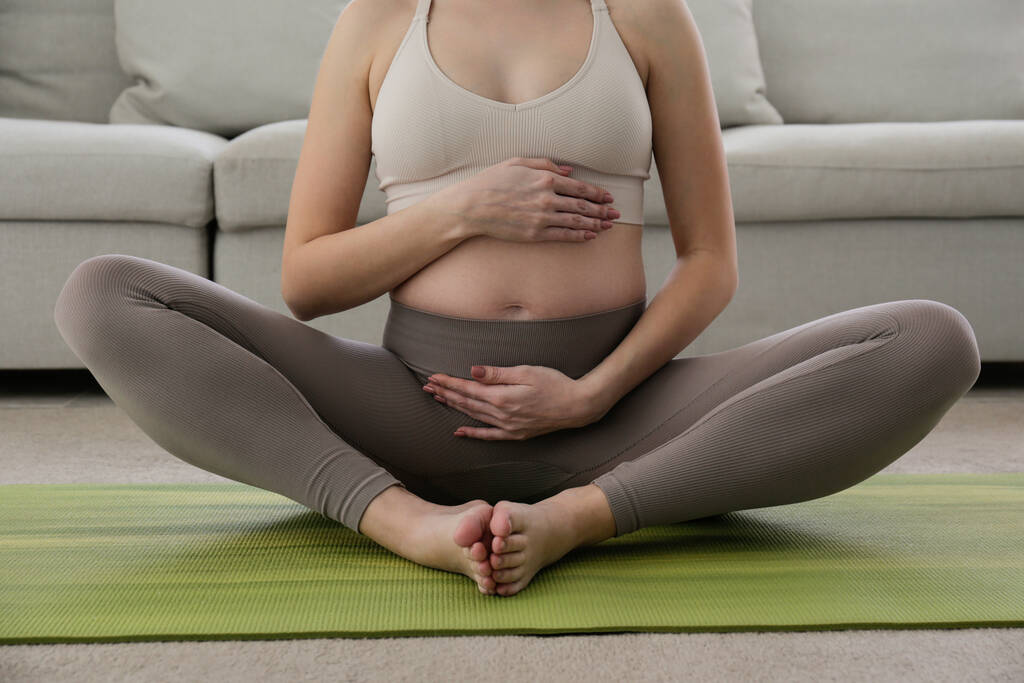 Νεαρή όμορφη γυναίκα στο δεύτερο τρίμηνο της εγκυμοσύνης κάνει γιόγκα. Κοντινό πλάνο της εγκύου γυναίκας που κάθεται σε ένα χαλί σε στάση λωτού και διαλογίζεται. Υγιής έννοια της εγκυμοσύνης. Ιστορικό, χώρος αντιγραφής. - Φωτογραφία, εικόνα