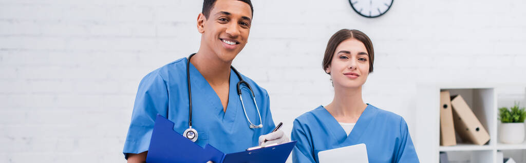 Cheerful interracial veterinarians with clipboard and digital tablet looking at camera in clinic, banner  - Photo, Image