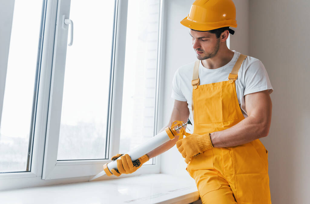 Handyman in yellow uniform works with glue for window indoors. House renovation conception. - 写真・画像