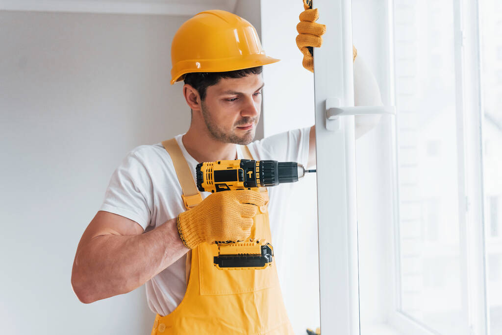Handyman en uniforme jaune installe une nouvelle fenêtre en utilisant un tournevis automatique. Conception de rénovation maison. - Photo, image