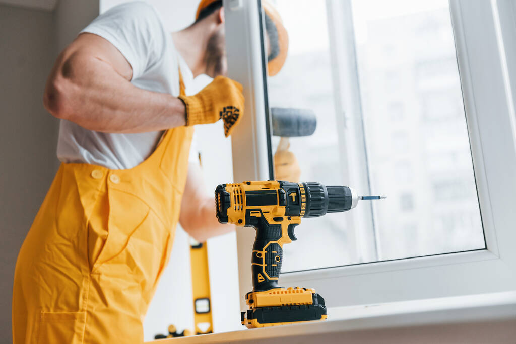 Handyman in yellow uniform installs new window by using automatic screwdriver. House renovation conception. - Foto, Imagen