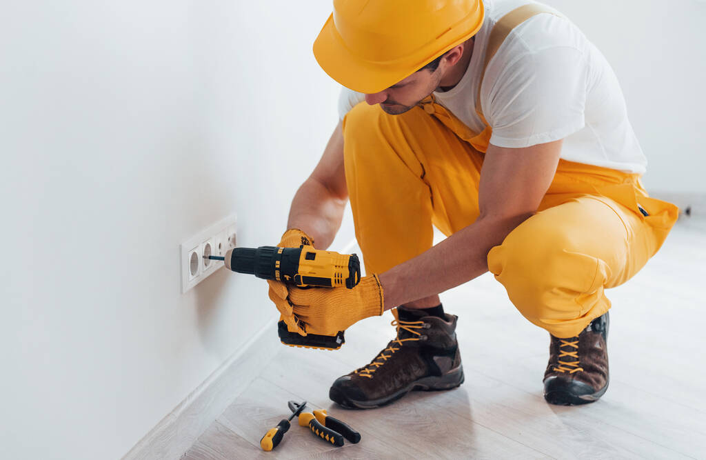 Handyman in geel uniform werkt met elektriciteit en het installeren van een nieuw stopcontact met behulp van automatische schroevendraaier. Conceptie woningrenovatie. - Foto, afbeelding
