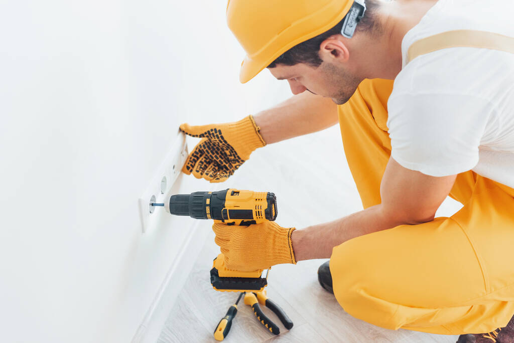 Handyman en uniforme amarillo trabaja con electricidad e instala un nuevo zócalo mediante el uso de un destornillador automático. Concepto de renovación de casas. - Foto, imagen