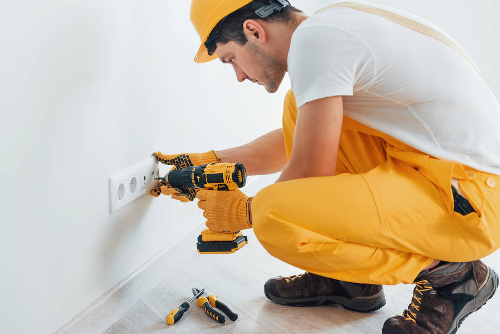 Handyman in yellow uniform works with electricity and installing new socket by using automatic screwdriver. House renovation conception. - Valokuva, kuva