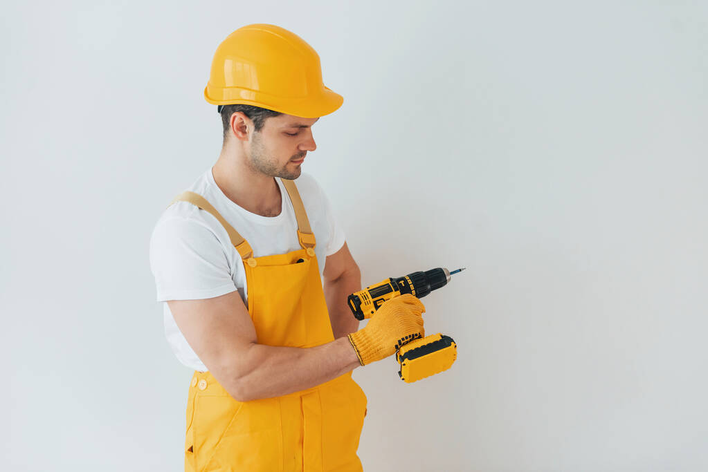 Handyman in geel uniform staande tegen witte muur met automatische schroevendraaier. Conceptie woningrenovatie. - Foto, afbeelding