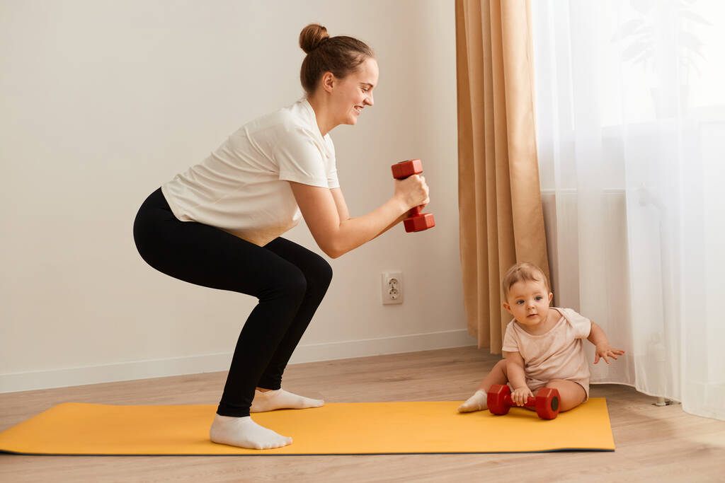 Side view portrait of slim athletic woman wearing white t shirt, standing on yoga mat, squatting with dumbbells in hands, training legs, butts and arms while looking after baby. - Foto, imagen