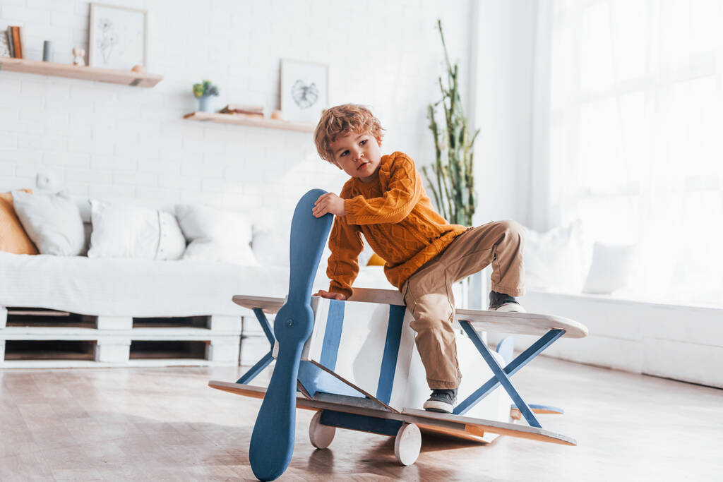 Мальчик в форме ретро-пилота веселится с игрушечным самолетом в помещении. - Фото, изображение